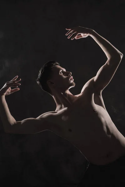 Un jeune danseur de ballet masculin avec des leggings noirs et un torse nu effectue des mouvements de danse sur un fond gris grunge, avec une lumière de lumières et de fumée . — Photo