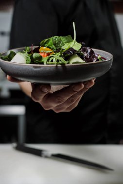 Siyah önlük Cook erkekte bir tabak salata elinde tutuyor. Mutfak restoranın arka planı.