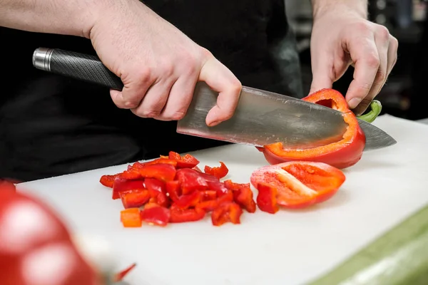 Chef maschio taglia verdure per insalata in un ristorante in un grembiule nero. Tagliere bianco, primo piano delle mani . — Foto Stock