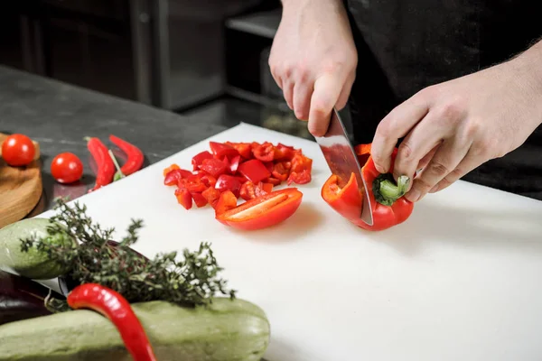 Chef maschio taglia verdure per insalata in un ristorante in un grembiule nero. Tagliere bianco, primo piano delle mani . — Foto Stock