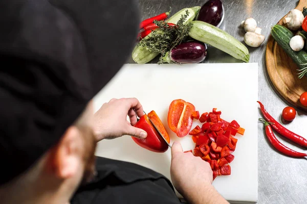 Chef maschio taglia verdure per insalata in un ristorante in un grembiule nero. Tagliere bianco, primo piano delle mani . — Foto Stock