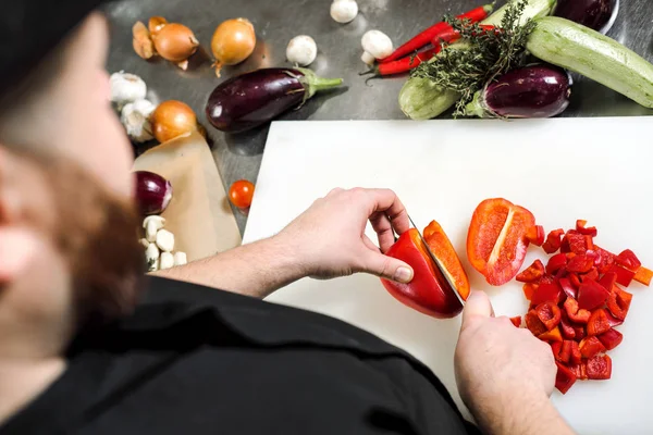 Chef maschio taglia verdure per insalata in un ristorante in un grembiule nero. Tagliere bianco, primo piano delle mani . — Foto Stock