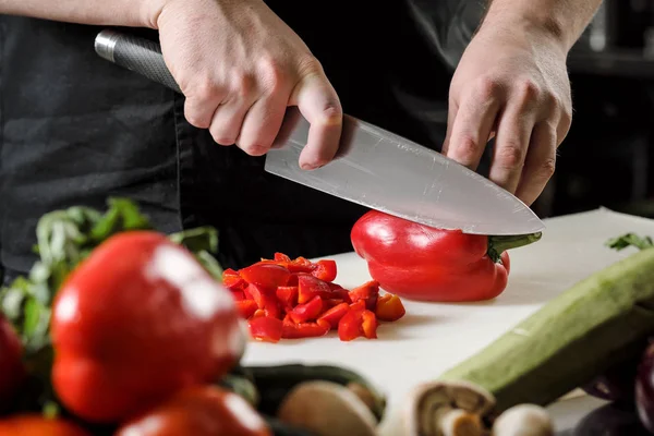 Chef maschio taglia verdure per insalata in un ristorante in un grembiule nero. Tagliere bianco, primo piano delle mani . — Foto Stock