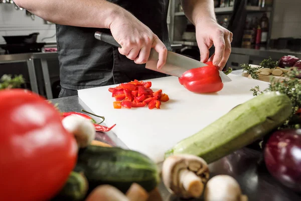 Chef maschio taglia verdure per insalata in un ristorante in un grembiule nero. Tagliere bianco, primo piano delle mani . — Foto Stock
