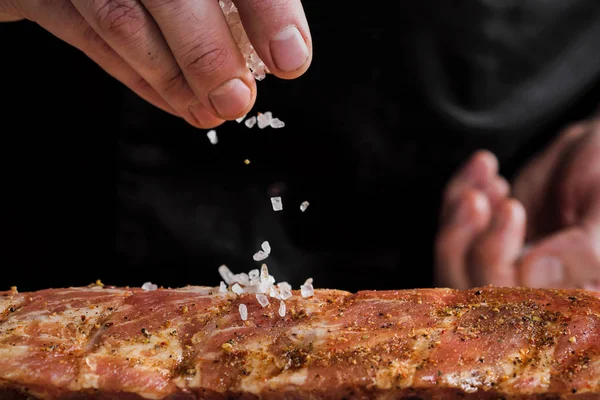Kawałek surowego mięsa, żeberka wołowe. Ręka szefa mężczyzna umieszcza sól i przyprawy na ciemnym tle. — Zdjęcie stockowe