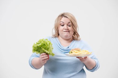 Yeşil salata yaprakları ve fast food, hamburger ve patates kızartması bir tabak holding beyaz bir arka plan üzerinde rahat mavi giysili genç kabarık şişman kadın. Doğru beslenme ve diyet.