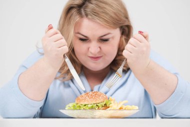 Beyaz bir arka planda hevesle fast food, hamburger ve patates kızartması yemek masasında rahat mavi giysili genç düzgün vücutlu şişman kadın. Doğru beslenme ve diyet.