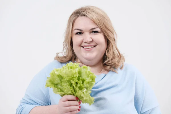 Egy fiatal buja kövér nő a gazdaság zöld saláta fehér háttér alkalmi kékruhás elhagyja a kezét és a száj. — Stock Fotó