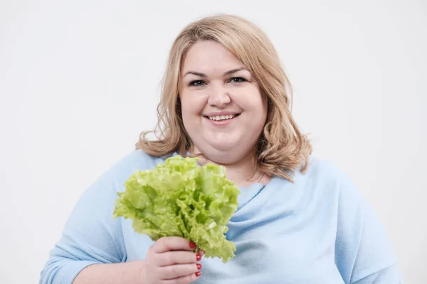 Een jonge weelderige dikke vrouw in casual blauwe kleding op een witte achtergrond met groene sla bladeren in haar handen en mond. — Stockfoto