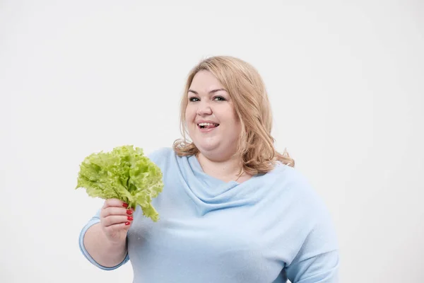 Молодая пышная толстая женщина в повседневной голубой одежде на белом фоне с зелеными листьями салата в руках и рту . — стоковое фото