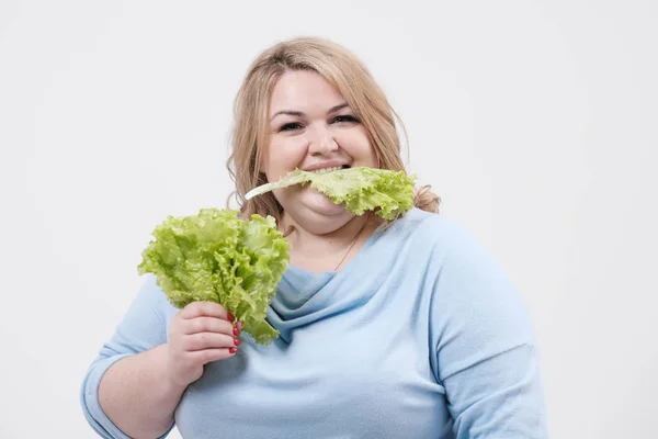 Молодая пышная толстая женщина в повседневной голубой одежде на белом фоне с зелеными листьями салата в руках и рту . — стоковое фото