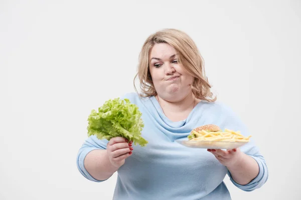 Молода пухнаста товста жінка в повсякденному синьому одязі на білому тлі, що тримає зелене листя салату і тарілку фаст-фуду, гамбургер і картопля фрі. Дієта і правильне харчування . — стокове фото