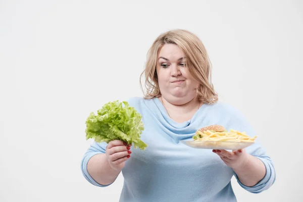 Mulher gorda fofa jovem em roupa azul casual em um fundo branco que mantém folhas de salada verdes e um prato de fast food, hambúrguer e batatas fritas. Dieta e nutrição adequada . — Fotografia de Stock