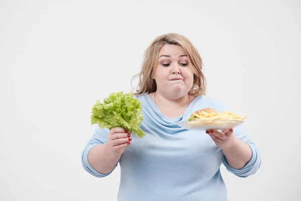 Молодая пушистая толстая женщина в повседневной голубой одежде на белом фоне с зелеными листьями салата и тарелкой фаст-фуда, гамбургером и картошкой фри. Диета и правильное питание . — стоковое фото