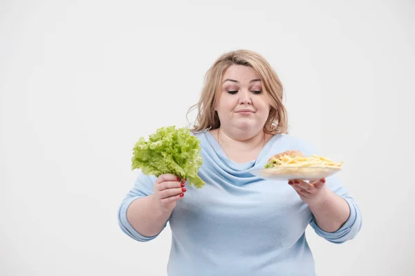 Молодая пушистая толстая женщина в повседневной голубой одежде на белом фоне с зелеными листьями салата и тарелкой фаст-фуда, гамбургером и картошкой фри. Диета и правильное питание . — стоковое фото