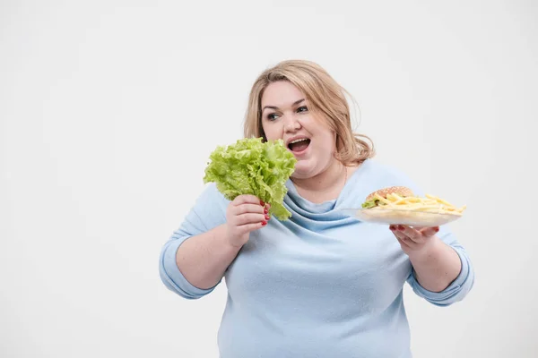 Jeune femme grasse moelleuse en vêtements bleus décontractés sur un fond blanc tenant des feuilles de salade vertes et une assiette de restauration rapide, hamburger et frites. Alimentation et bonne nutrition . — Photo