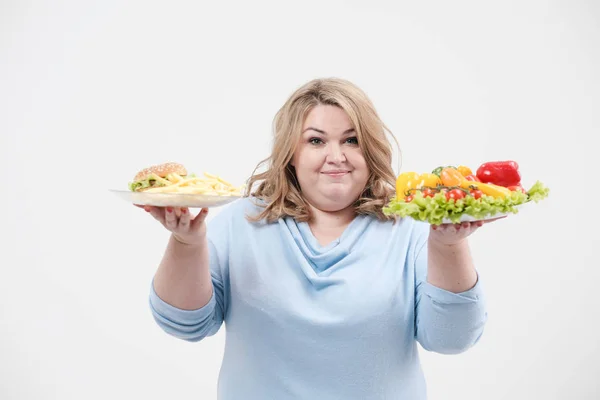Jeune femme de graisse luxuriante dans des vêtements bleus décontractés sur un fond blanc tenant une salade de légumes et une assiette de restauration rapide, hamburger et frites. Alimentation et bonne nutrition . — Photo