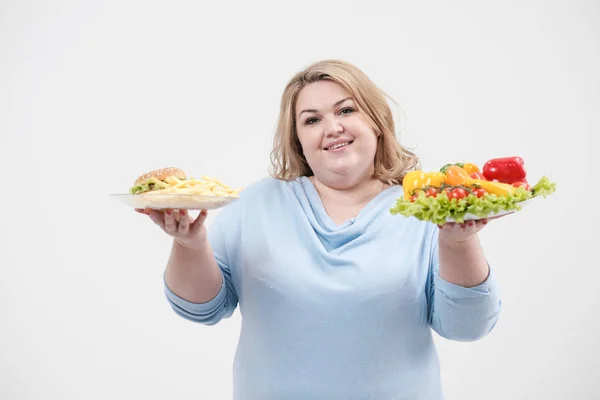 Jeune femme de graisse luxuriante dans des vêtements bleus décontractés sur un fond blanc tenant une salade de légumes et une assiette de restauration rapide, hamburger et frites. Alimentation et bonne nutrition . — Photo