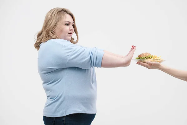 Una mujer gorda magnífica joven en ropa azul casual sobre un fondo blanco se niega a la comida rápida que se le ofrece, una hamburguesa y papas fritas. Dieta y nutrición adecuada . — Foto de Stock