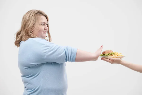 Una mujer gorda magnífica joven en ropa azul casual sobre un fondo blanco se niega a la comida rápida que se le ofrece, una hamburguesa y papas fritas. Dieta y nutrición adecuada . — Foto de Stock