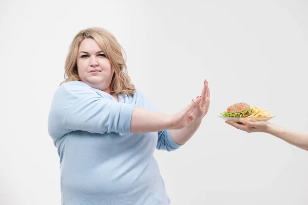 Una mujer gorda magnífica joven en ropa azul casual sobre un fondo blanco se niega a la comida rápida que se le ofrece, una hamburguesa y papas fritas. Dieta y nutrición adecuada . — Foto de Stock