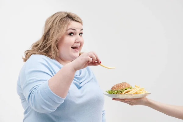Молодая пышная толстая женщина в повседневной голубой одежде на белом фоне, крадущаяся с тарелки гамбургера и картошки фри. Диета и правильное питание . — стоковое фото