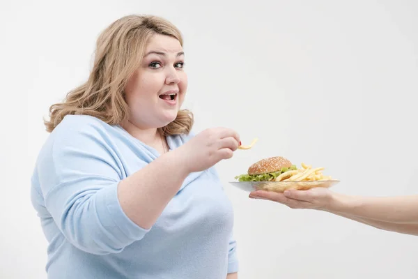 Joven mujer gorda con curvas en ropa azul casual sobre un fondo blanco que se escabulle de un plato de hamburguesa y papas fritas. Dieta y nutrición adecuada . — Foto de Stock