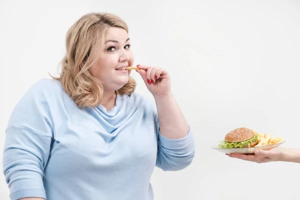 Młoda kobieta krzywego tłuszczu w błękitne ubranie na białym tle skradanie się płytkę z hamburger i frytki. Prawidłowe odżywianie i dieta. — Zdjęcie stockowe