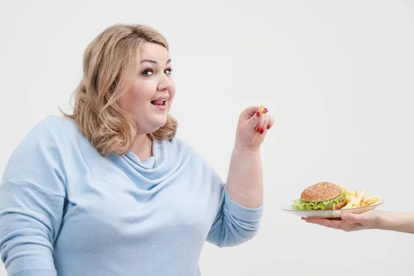 Jovem mulher gorda curvilínea em roupas azuis casuais em um fundo branco saindo sorrateiramente de um prato de hambúrguer e batatas fritas. Dieta e nutrição adequada . — Fotografia de Stock