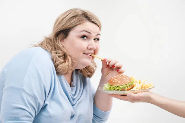 Jovem mulher gorda curvilínea em roupas azuis casuais em um fundo branco saindo sorrateiramente de um prato de hambúrguer e batatas fritas. Dieta e nutrição adequada . — Fotografia de Stock