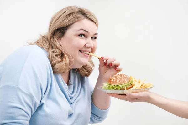 Joven mujer gorda con curvas en ropa azul casual sobre un fondo blanco que se escabulle de un plato de hamburguesa y papas fritas. Dieta y nutrición adecuada . — Foto de Stock