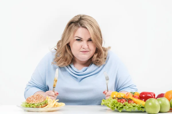 Молодая пышная толстая женщина в повседневной голубой одежде на белом фоне за столом выбирает, что есть: фаст-фуд или здоровое питание. Диета и правильное питание . — стоковое фото