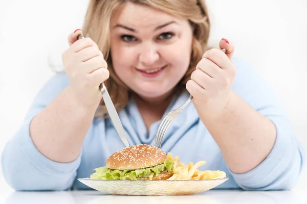 Молодая пышная толстая женщина в повседневной голубой одежде на белом фоне за столом охотно ест фаст-фуд, гамбургер и картошку фри. Диета и правильное питание . — стоковое фото