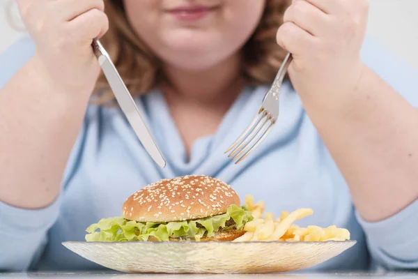 Jeune femme obèse courbée en vêtements bleus décontractés sur un fond blanc à la table mangeant avec impatience de la restauration rapide, hamburger et frites. Alimentation et bonne nutrition . — Photo