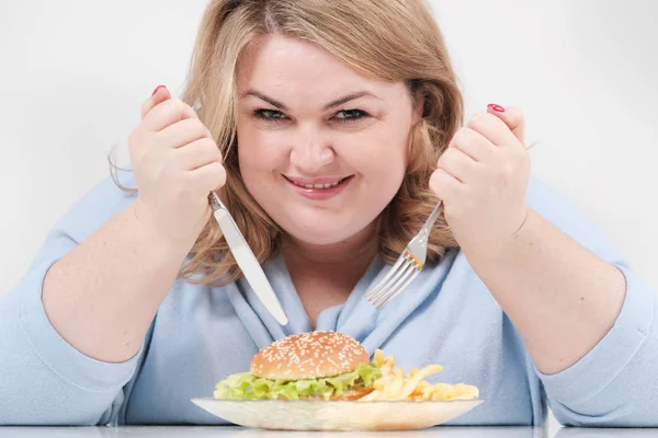 Молодая пышная толстая женщина в повседневной голубой одежде на белом фоне за столом охотно ест фаст-фуд, гамбургер и картошку фри. Диета и правильное питание . — стоковое фото