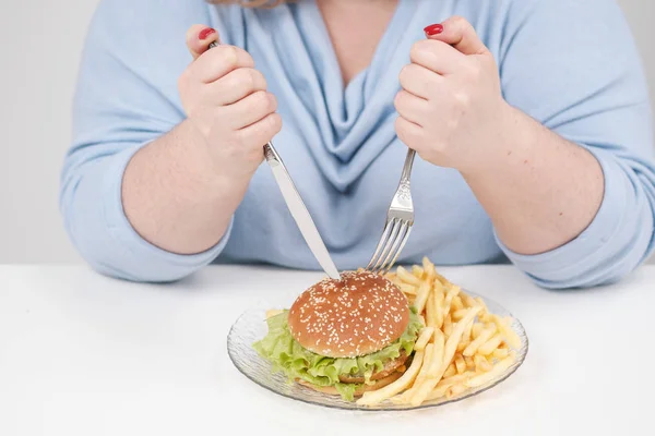 Joven mujer gorda con curvas en ropa azul casual sobre un fondo blanco en la mesa comiendo con entusiasmo comida rápida, hamburguesa y papas fritas. Dieta y nutrición adecuada . — Foto de Stock