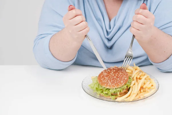 Joven mujer gorda con curvas en ropa azul casual sobre un fondo blanco en la mesa comiendo con entusiasmo comida rápida, hamburguesa y papas fritas. Dieta y nutrición adecuada . — Foto de Stock