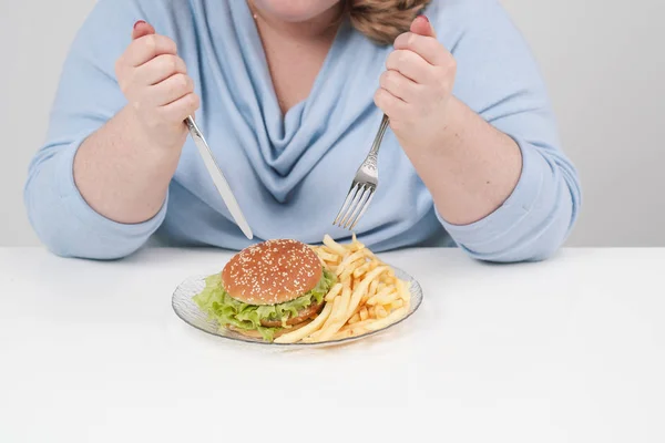 Молода пишна товста жінка в повсякденному блакитному одязі на білому тлі за столом з нетерпінням їсть фастфуд, гамбургер і картоплю фрі. Дієта і правильне харчування . — стокове фото