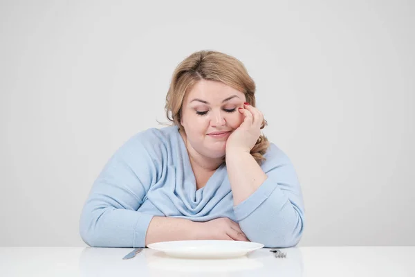 Молодая пышная толстая женщина в повседневной голубой одежде на белом фоне за столом сидит перед пустой тарелкой и голодает. Диета и правильное питание . — стоковое фото