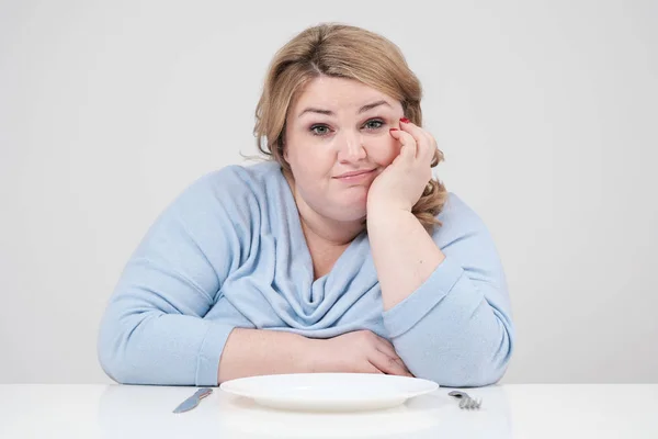 Молодая пышная толстая женщина в повседневной голубой одежде на белом фоне за столом сидит перед пустой тарелкой и голодает. Диета и правильное питание . — стоковое фото