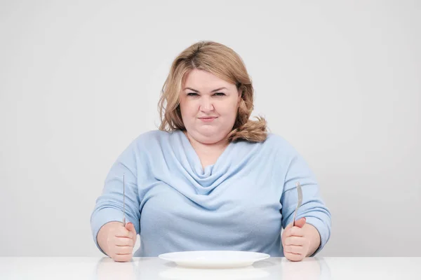 Jonge bochtige vet vrouw in casual blauwe kleding op een witte achtergrond aan de tafel zitten voor een leeg bord en honger. Voeding en de juiste voeding. — Stockfoto