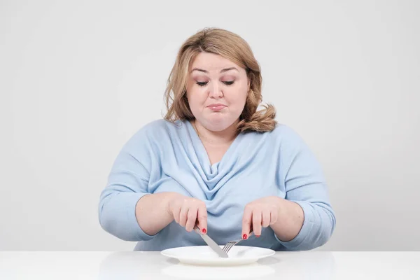 Genç curvy masada boş bir levha önünde oturan ve açlıktan beyaz bir arka plan üzerinde rahat mavi giysili kadın şişman. Doğru beslenme ve diyet.