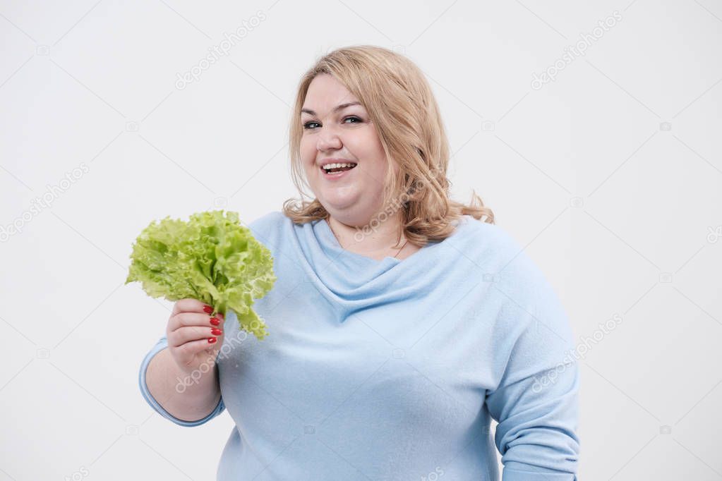 A young lush fat woman in casual blue clothes on a white background holding green lettuce leaves in her hands and mouth.