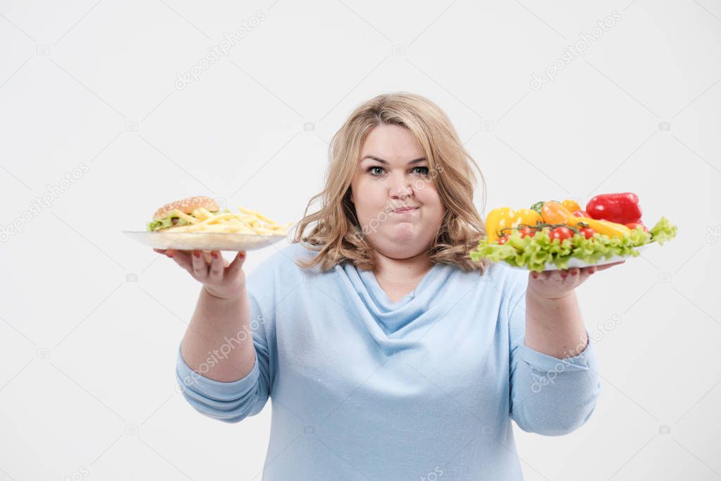Young lush fat woman in casual blue clothes on a white background holding a vegetable salad and a plate of fast food, hamburger and fries. Diet and proper nutrition.