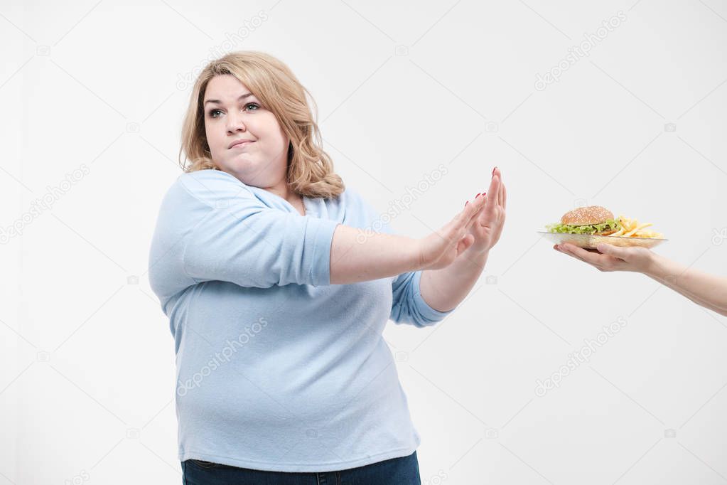 A young magnificent fat woman in casual blue clothes on a white background refuses from the fast food offered to her, a hamburger and french fries. Diet and proper nutrition.