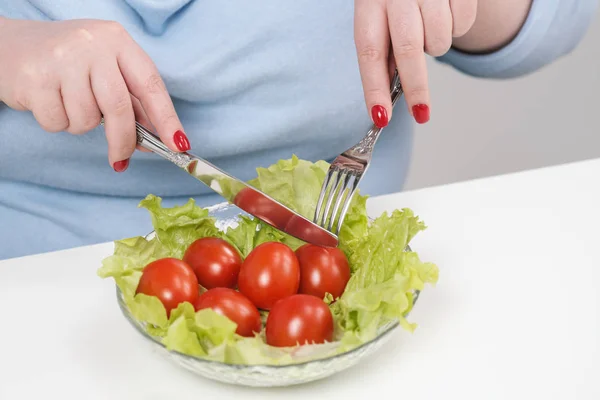 Mladá svěží tlustá žena v neformálním modré oblečení na bílém pozadí u stolu a jí zeleninový salát s rajčaty. Diety a zdravá výživa. — Stock fotografie