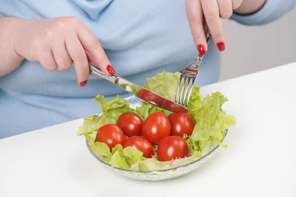 Jeune femme riche en graisse dans des vêtements bleus décontractés sur un fond blanc à la table et mange une salade de légumes avec des tomates. Alimentation et bonne nutrition . — Photo