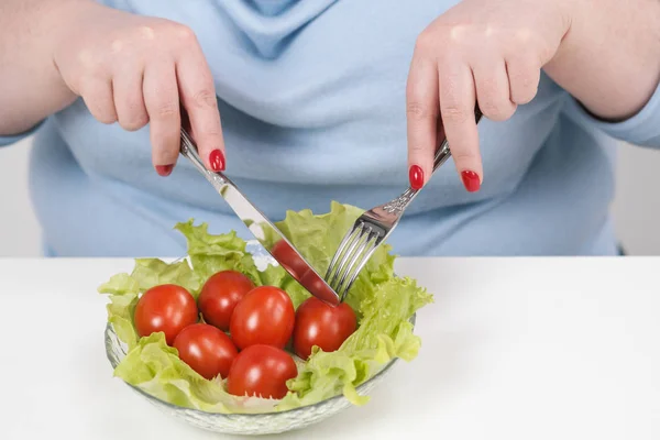 Jovem mulher gorda exuberante em roupas azuis casuais em um fundo branco na mesa e come uma salada de legumes com tomates. Dieta e nutrição adequada . — Fotografia de Stock