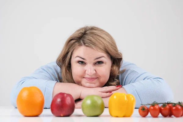 Молодая пышная толстая женщина в повседневной голубой одежде на белом фоне за столом, перед ней в ряд выкладывали овощи и фрукты. Диета и правильное питание . — стоковое фото