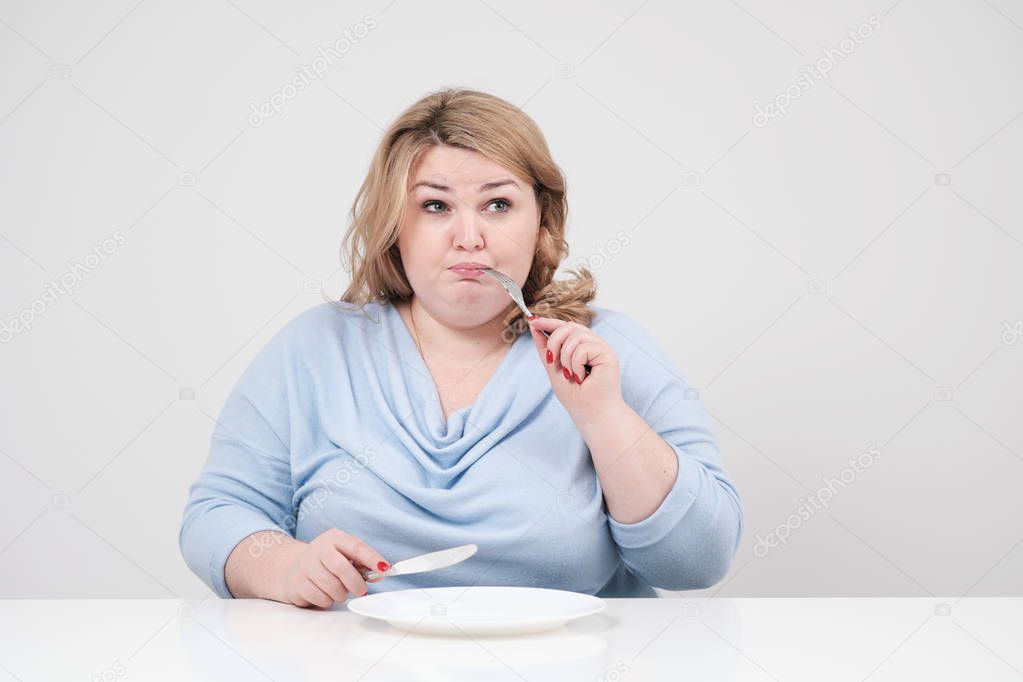 Young curvy fat woman in casual blue clothes on a white background at the table sitting in front of an empty plate and starving. Diet and proper nutrition.